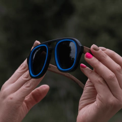manos de mujer sosteniendo anteojos de sol de madera (patillas) y frente de acetato negro con aro incrustado color azul, con lentes grises modelo Hanoli BOLD estilo aviador marca Nómade